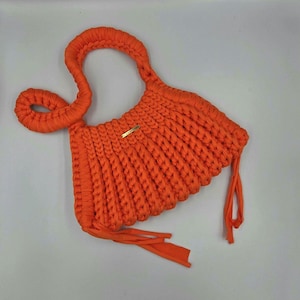 May include: Bright orange knitted handbag with a braided handle and decorative tassels. The bag features a small gold-colored rectangular tag. The bag is made of thick, chunky knit material.