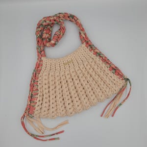 May include: A cream-colored, handmade crochet bag with a braided strap and floral trim. The bag features a scalloped edge and decorative tassels. The strap and trim incorporate shades of pink, green, and cream.