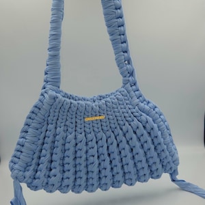 May include: A light blue, handmade crochet bag with a rounded shape and braided handles. The bag features a gold-colored rectangular tag with text. The bag is made of a soft, textured material and has a simple, elegant design.