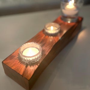Könnte beinhalten: Ein hölzerner Kerzenhalter mit drei durchsichtigen Glas-Teelichthaltern. Jedes Teelicht enthält eine brennende weiße Kerze. Der Holzsockel ist rechteckig und hat eine natürliche Holzmaserung.