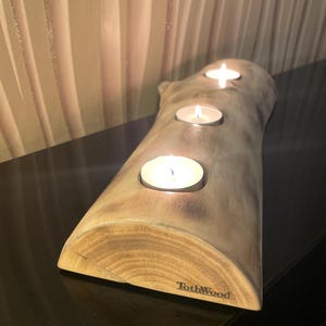 Könnte beinhalten: Ein hölzerner Kerzenhalter mit drei brennenden Teelichtern. Der Halter besteht aus hellem, strukturiertem Holz und trägt den Text "TothWood" auf der Seite. Die Kerzen befinden sich in der Mitte des Holzes.