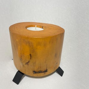 Könnte beinhalten: Ein Kerzenhalter aus Holz mit einer natürlichen Holzoptik und schwarzen Metallbeinen. Der Halter hat eine kleine, runde Öffnung für ein Teelicht. Der Text "Truly Wood" ist in das Holz gebrannt.