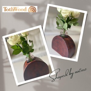 Könnte beinhalten: Eine dunkelbraune Holzvase in halbkreisförmiger Form, die weiße Rosen zeigt. Die Vase hat einen kleinen Glaseinsatz für die Blumen. Der Text "Shaped by nature" ist in Kursivschrift auf dem Bild zu sehen.