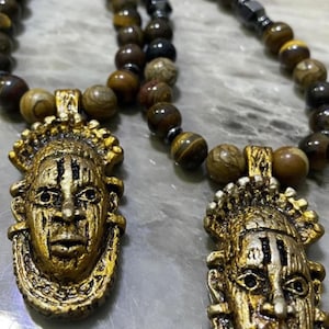 May include: Two gold-toned pendants with African tribal mask designs hang from a brown and black beaded necklace. The necklace is made of tiger's eye and hematite beads.