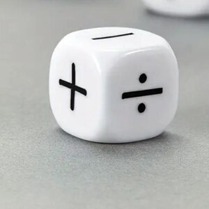 May include: A white die with black symbols for addition, subtraction, and division. The die is sitting on a gray surface. The die is a cube shape.