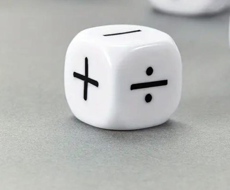 May include: A white die with black symbols for addition, subtraction, and division. The die is sitting on a gray surface. The die is a cube shape.