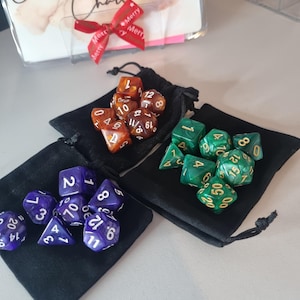 May include: Three sets of polyhedral dice in black drawstring bags. One set is purple with white numbers, another is green with gold numbers, and the third is brown with white numbers. A decorative card with a red ribbon is in the background.
