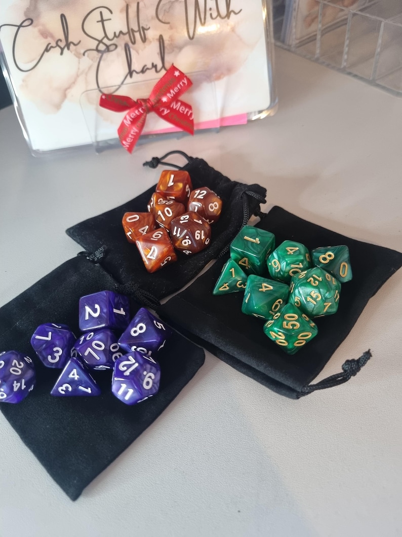 May include: Three sets of polyhedral dice in black drawstring bags. One set is purple with white numbers, another is green with gold numbers, and the third is brown with white numbers. A decorative card with a red ribbon is in the background.