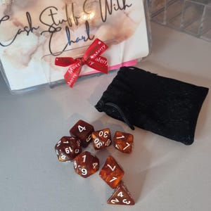 May include: A collection of seven brown and orange dice with white numbering, next to a black pouch. A clear plastic holder with the text "Cash Stuff With Charl" and a red ribbon with "Merry" text is also visible.