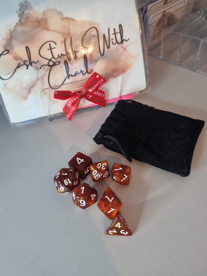 May include: A collection of seven brown and orange dice with white numbering, next to a black pouch. A clear plastic holder with the text "Cash Stuff With Charl" and a red ribbon with "Merry" text is also visible.