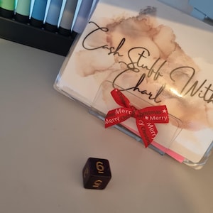 May include: A purple die with gold numbering, displaying the number 6. A clear plastic cover with the words "Cash Stuff With Charl" is visible, along with a red ribbon that says "Merry Christmas". A set of colorful pens are in the background.