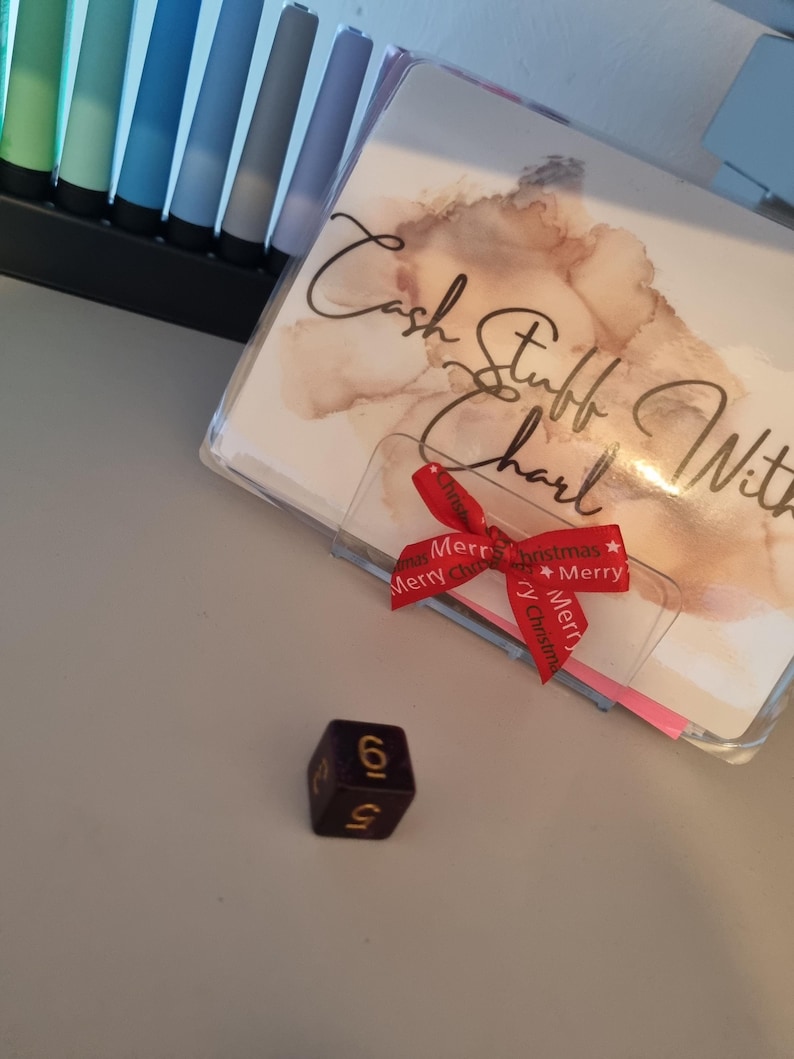 May include: A purple die with gold numbering, displaying the number 6. A clear plastic cover with the words "Cash Stuff With Charl" is visible, along with a red ribbon that says "Merry Christmas". A set of colorful pens are in the background.