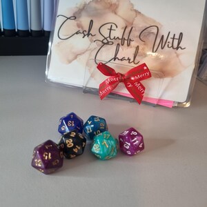 May include: A collection of polyhedral dice in various colors, including purple, blue, and teal, with white and gold numbering. A clear plastic holder with the text "Cash Stuff With Charl" and a red ribbon with "Merry" text is also present.