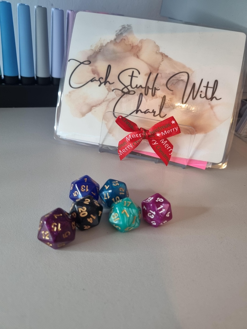May include: A collection of polyhedral dice in various colors, including purple, blue, and teal, with white and gold numbering. A clear plastic holder with the text "Cash Stuff With Charl" and a red ribbon with "Merry" text is also present.
