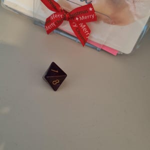 May include: A dark purple, eight-sided die with gold numbering, displaying the number 1. A red ribbon with "Merry Christmas" text is tied to a clear plastic holder with a card inside. The card has handwritten text.