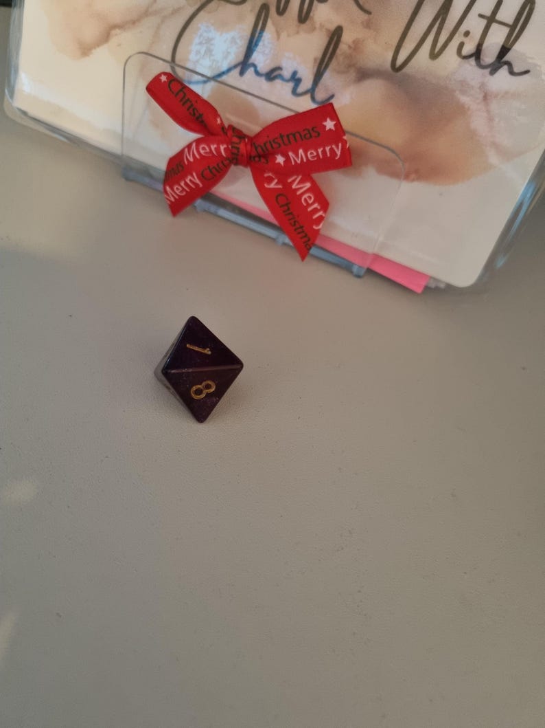 May include: A dark purple, eight-sided die with gold numbering, displaying the number 1. A red ribbon with "Merry Christmas" text is tied to a clear plastic holder with a card inside. The card has handwritten text.