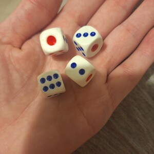 May include: Four white dice with blue and red markings are held in an open palm. The dice have blue dots and red dots, with varying numbers of dots on each die. The dice are small and appear to be made of plastic.