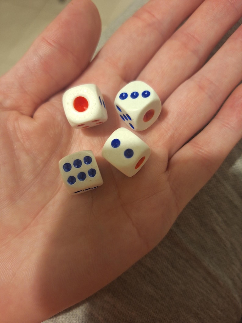 May include: Four white dice with blue and red markings are held in an open palm. The dice have blue dots and red dots, with varying numbers of dots on each die. The dice are small and appear to be made of plastic.