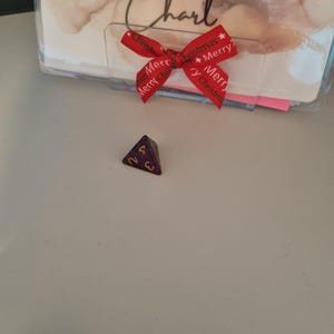 May include: A purple and gold four-sided die with the number 2 and 4 visible. A red ribbon with the words "Merry Christmas" is tied in a bow. The text "Cash Stuff With Charl" is visible in the background.