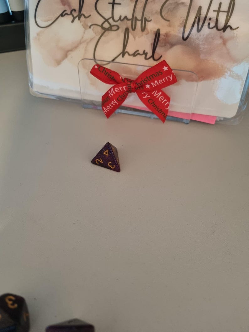 May include: A purple and gold four-sided die with the number 2 and 4 visible. A red ribbon with the words "Merry Christmas" is tied in a bow. The text "Cash Stuff With Charl" is visible in the background.