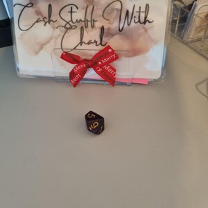 May include: A black and gold six-sided die with the numbers 5 and 6 visible. In the background is a clear plastic holder with the text "Cash Stuff With Charl" and a red ribbon with "Merry Christmas" printed on it.