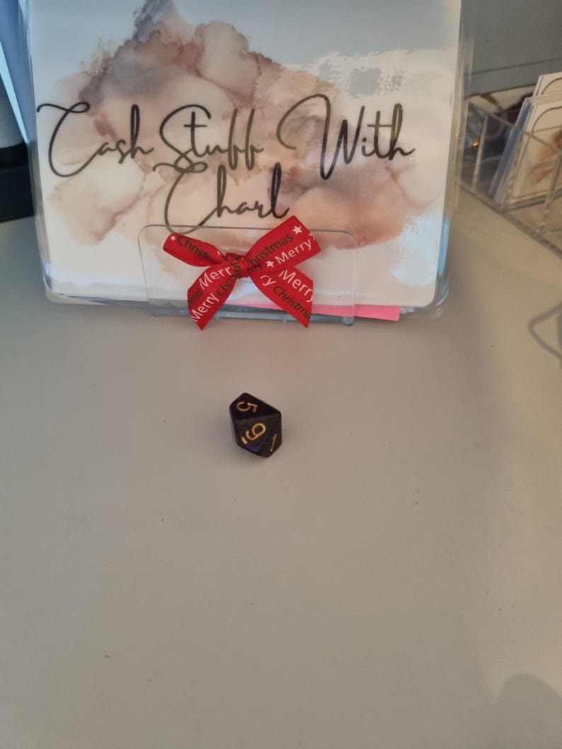 May include: A black and gold six-sided die with the numbers 5 and 6 visible. In the background is a clear plastic holder with the text "Cash Stuff With Charl" and a red ribbon with "Merry Christmas" printed on it.