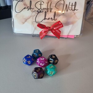May include: Six multi-sided dice in various colors, including blue, purple, and green, are arranged on a light gray surface. A clear plastic holder with the text "Cash Stuff With Charl" and a red ribbon with "Merry" text are in the background.