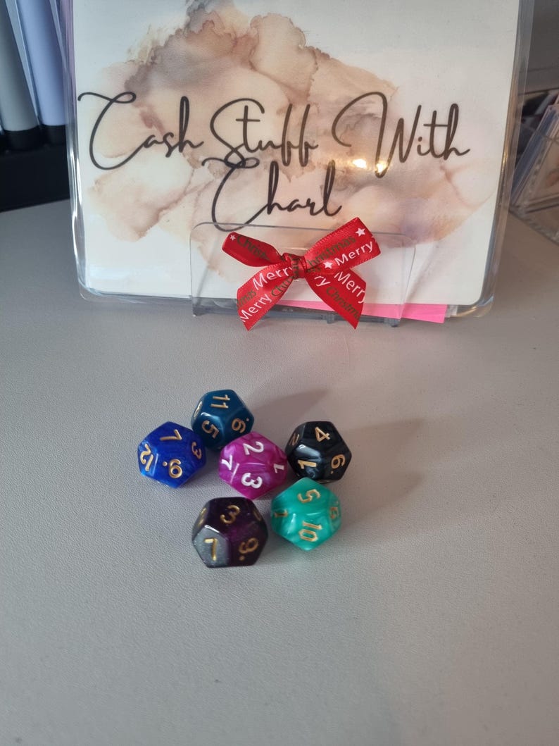 May include: Six multi-sided dice in various colors, including blue, purple, and green, are arranged on a light gray surface. A clear plastic holder with the text "Cash Stuff With Charl" and a red ribbon with "Merry" text are in the background.