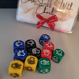 May include: A collection of colorful dice featuring hand gestures for the game Rock, Paper, Scissors. The dice are in various colors, including blue, black, red, yellow, and green. A clear plastic holder with text and a red ribbon is in the background.