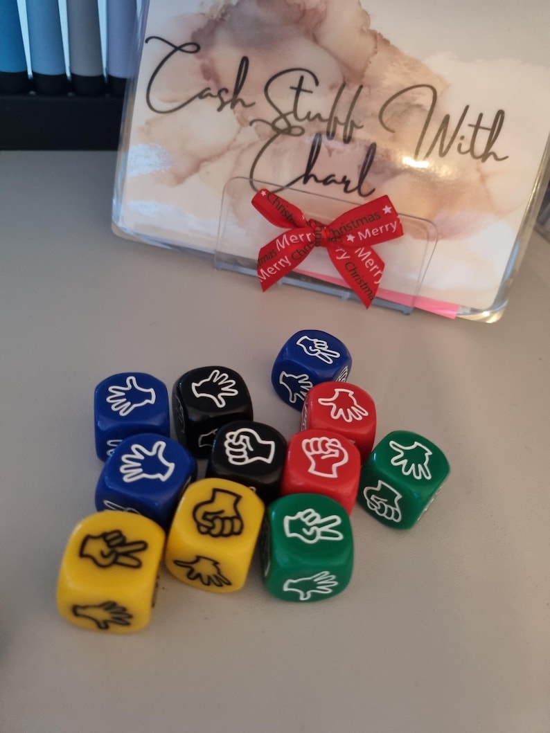 May include: A collection of colorful dice featuring hand gestures for the game Rock, Paper, Scissors. The dice are in various colors, including blue, black, red, yellow, and green. A clear plastic holder with text and a red ribbon is in the background.