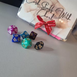 May include: A collection of polyhedral dice in various colors, including purple, blue, green, and black, with gold numbering. A clear plastic sleeve with the text "Cash Stuff With Charl" is tied with a red ribbon.