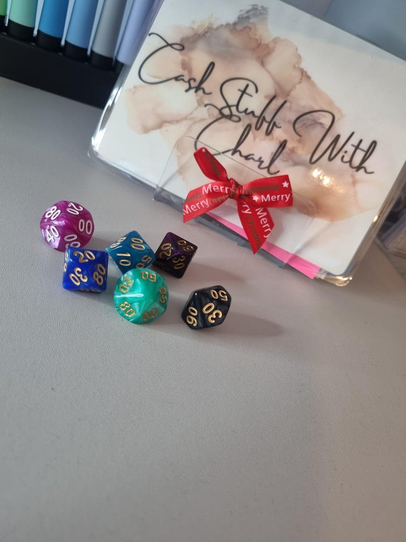 May include: A collection of polyhedral dice in various colors, including purple, blue, green, and black, with gold numbering. A clear plastic sleeve with the text "Cash Stuff With Charl" is tied with a red ribbon.