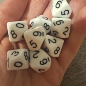 May include: A collection of white dice with black numbers, including six-sided and multi-sided dice. The dice are held in a hand, showcasing the variety of shapes and numbers for tabletop gaming.