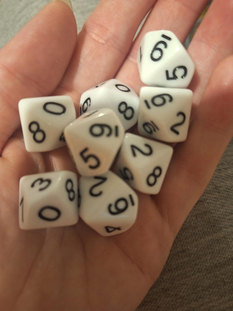 May include: A collection of white dice with black numbers, including six-sided and multi-sided dice. The dice are held in a hand, showcasing the variety of shapes and numbers for tabletop gaming.
