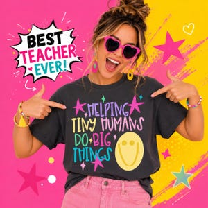 May include: A charcoal gray t-shirt featuring the text "HELPING TINY HUMANS DO BIG THINGS" in colorful lettering, accompanied by a yellow smiley face and star graphics. A speech bubble in the corner displays the text "BEST TEACHER EVER!"