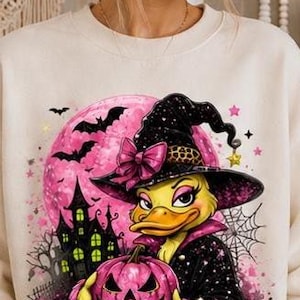 Op de afbeelding: Een crèmekleurig sweatshirt met ronde hals en een grafische print van een gele eend verkleed als heks, met een zwarte hoed met een roze strik. De eend houdt een roze pompoen vast voor een grote roze maan, een donker kasteel en vleermuizen.