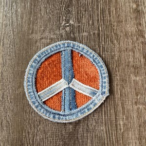 Puede incluir: Un parche redondo con el símbolo de la paz con un borde azul vaquero y detalles blancos y azules. El centro del parche es naranja. El parche probablemente está hecho de tela y está diseñado para ser cosido o planchado en ropa o accesorios.