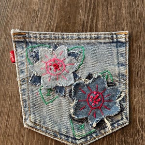 May include: A light blue denim pocket with two embroidered flower appliqués. The flowers have pink and red centers, with green stitched stems and leaves. A red tag is visible on the left side.