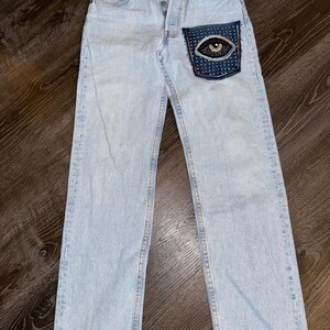 Op de afbeelding: Lichtblauwe denim jeans met een decoratieve patch op de achterzak. De patch heeft een oogontwerp met kleurrijk borduurwerk en kralenwerk. De jeans heeft een knoopsluiting en een klassieke vijf-zakken-stijl.