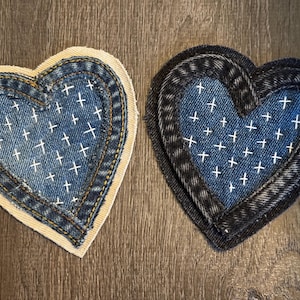 Könnte beinhalten: Zwei herzförmige Denim-Patches. Einer hat eine hellblaue Denim-Mitte mit weißen Kreuzstichdetails und einen beigen Rand. Der andere hat eine dunkelblaue Denim-Mitte mit weißen Kreuzstichdetails und einen dunkelblauen Rand.