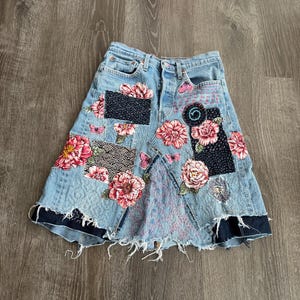 May include: Light blue denim skirt with floral and patchwork embellishments. The skirt features pink and red floral appliqués, patches of dark blue fabric, and frayed edges. The design includes butterfly and wave patterns.