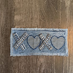 May include: A denim patch featuring the letters "XOXO" with heart shapes. The letters and hearts are made of a textured fabric, and the patch has a frayed edge. The patch is on a wooden surface.