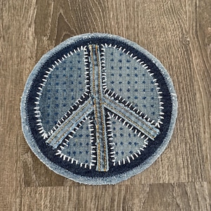 May include: A round denim patch featuring a peace symbol. The patch is made of blue denim with white stitching outlining the peace sign and the circular border. The background is a wooden surface.