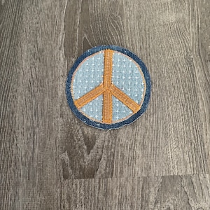 May include: A circular patch featuring a peace symbol. The patch has a light blue background with a white cross pattern, a brown peace symbol, and a dark blue denim border.