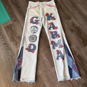 May include: Light-wash denim jeans featuring patchwork side panels with pink stitching and floral embroidery. The front displays appliquéd letters spelling GOOD KARMA, a peace sign, and an eye motif, all accented with small pink and red floral details.