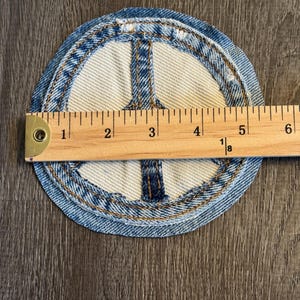May include: Circular denim patch featuring a peace sign design. The patch is approximately 4 inches in diameter. The peace sign is made of white fabric with blue denim stitching around the edges and the peace symbol.