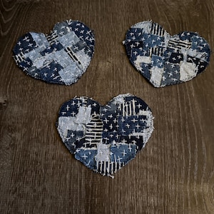 May include: Three heart-shaped denim patches with a patchwork design. Each heart is made of various denim pieces stitched together with white thread, creating a textured effect. The hearts are arranged on a dark wooden surface.