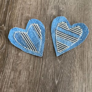 May include: Two heart-shaped denim patches with a blue and white striped fabric insert. The patches are made of blue denim and are stitched around the edges. They are placed on a wooden surface.