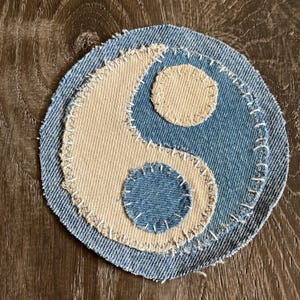 Puede incluir: Un parche circular con el símbolo del yin y el yang. El diseño está hecho de tela vaquera y beige, con costuras blancas que delinean las formas. El parche tiene un aspecto artesanal y desgastado.