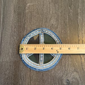 May include: A denim peace sign patch with a camouflage center, approximately 4 inches in diameter. The patch is made of blue denim with a light blue stitched border. A wooden ruler is placed across the patch for measurement.